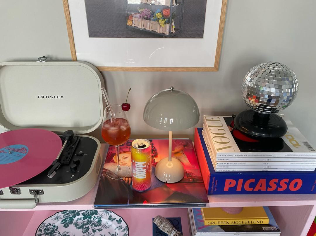 A vibrant setup featuring a Crosley turntable playing a pink vinyl, a stylish cocktail, and a can of häppi prebiotic soda, surrounded by art books and a retro disco ball for a perfect cozy vibe.