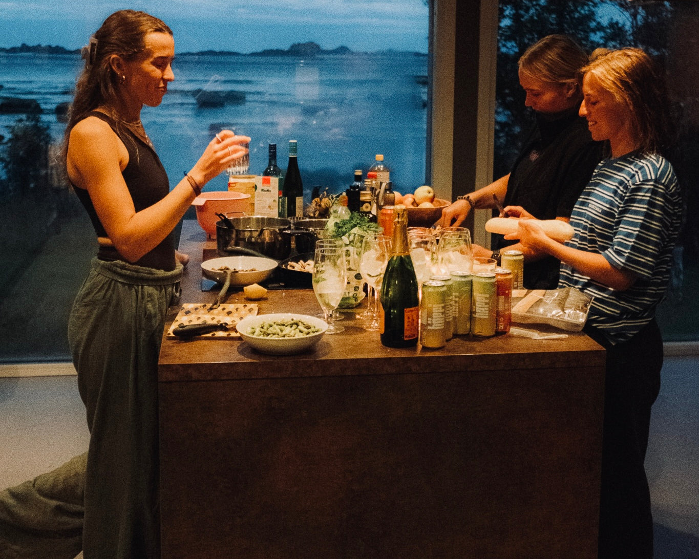 A cozy evening gathering with friends enjoying prebiotic hÄppi soda, surrounded by fresh ingredients, wellness drinks, and a stunning seaside view.