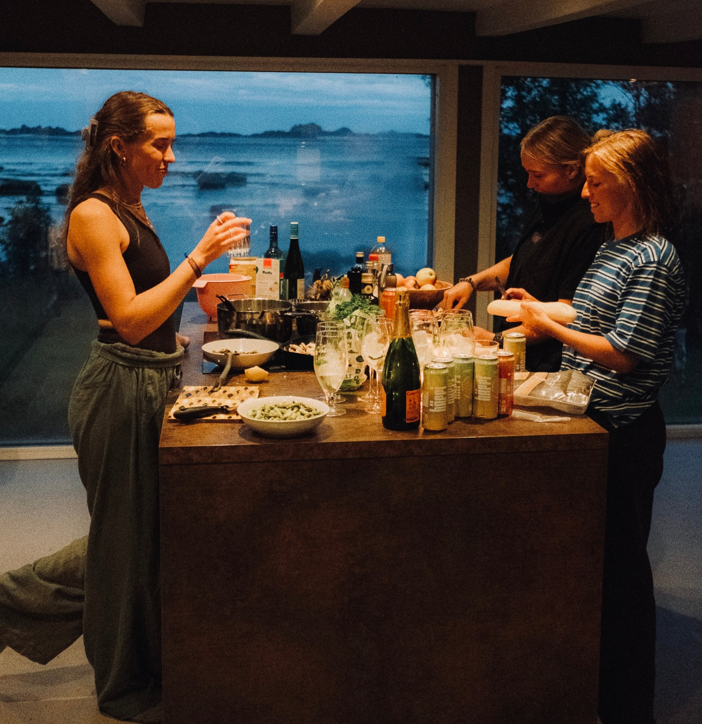 A cozy evening gathering with friends enjoying prebiotic hÄppi soda, surrounded by fresh ingredients, wellness drinks, and a stunning seaside view.