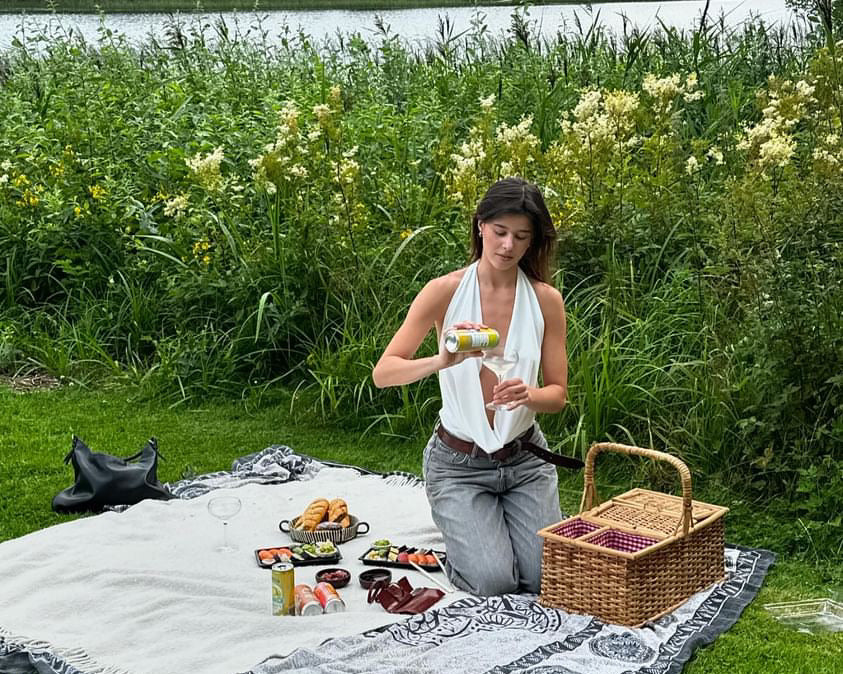 A beautiful woman enjoys a sunny picnic, pouring hÄppi prebiotic soda into a glass surrounded by flowers and healthy snacks. Perfect for outdoor gatherings, hÄppi sodas add a refreshing and gut-friendly boost to your picnic experience.