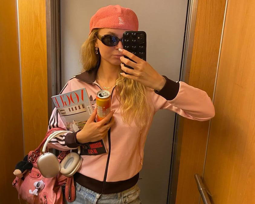 Young woman in a stylish pink jacket and hat taking a selfie in an elevator, holding a can of hÄppi prebiotic soda. She carries a pink bag and a magazine, showcasing a trendy and health-conscious lifestyle on the go.