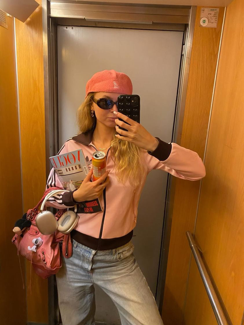 Young woman in a stylish pink jacket and hat taking a selfie in an elevator, holding a can of hÄppi prebiotic soda. She carries a pink bag and a magazine, showcasing a trendy and health-conscious lifestyle on the go.