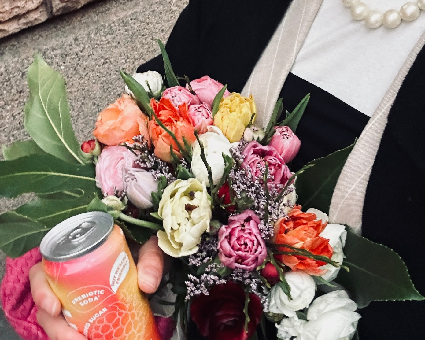 a person holding a colorful bouquet and a can of hÄppi raspberry prebiotic soda. The vibrant pink and orange can complements the bright flowers, creating a fresh, cheerful vibe. The stylish black coat and pearl necklace add an elegant touch.