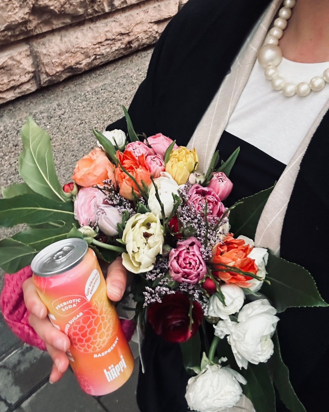 a person holding a colorful bouquet and a can of hÄppi raspberry prebiotic soda. The vibrant pink and orange can complements the bright flowers, creating a fresh, cheerful vibe. The stylish black coat and pearl necklace add an elegant touch.