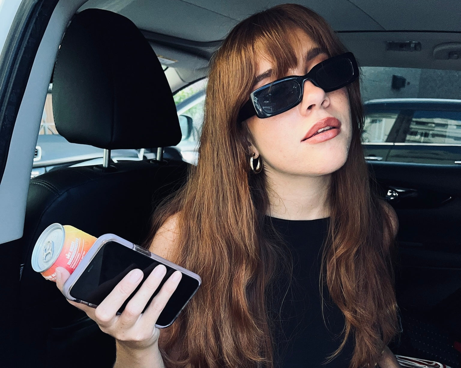 A woman with long brown hair, wearing black sunglasses and an all-black outfit, sits in a car holding a can of drink and a smartphone. The image evokes a stylish, casual vibe 