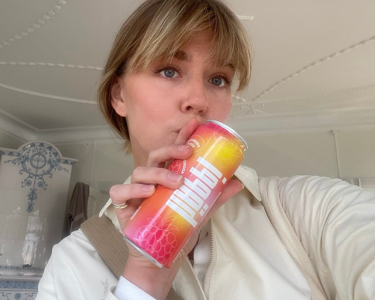 A woman enjoys a häppi prebiotic soda, capturing a refreshing and stylish moment indoors with a bright, fruity drink in hand.