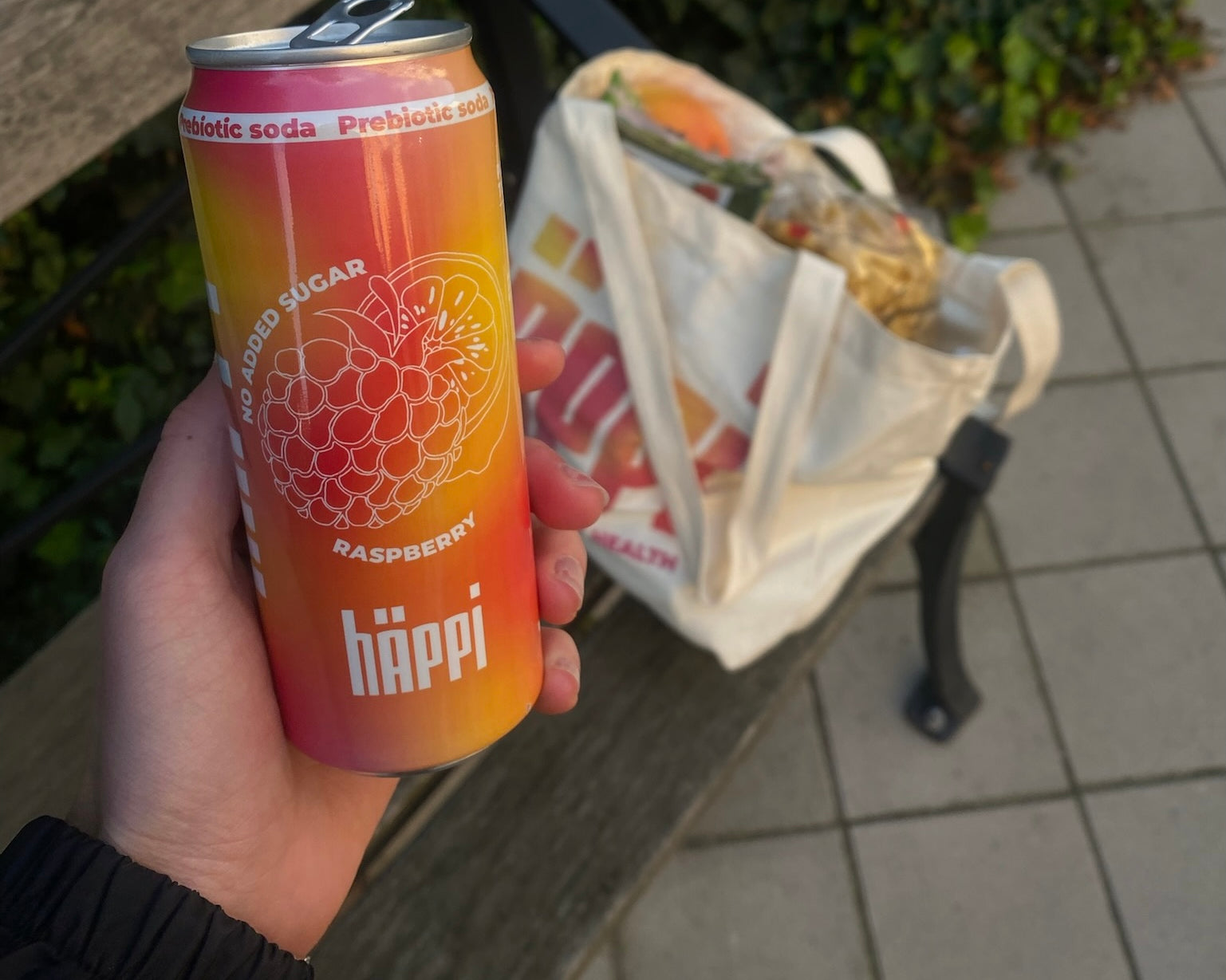 A vibrant can of hÄppi prebiotic soda in raspberry flavor held in hand, showcasing "No Added Sugar" labeling. Positioned outdoors on a park bench, with a reusable tote bag filled with groceries in the background.