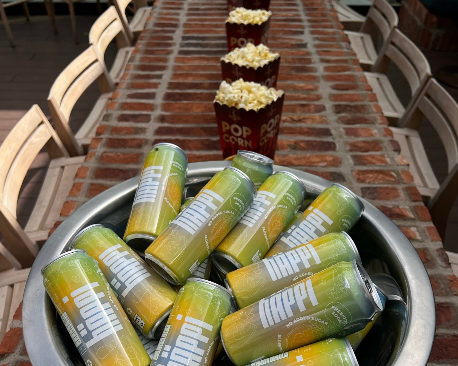 A bowl filled with cans of häppi prebiotic soda is placed on a long brick table, lined with popcorn buckets, creating a setup perfect for a movie night or casual gathering.