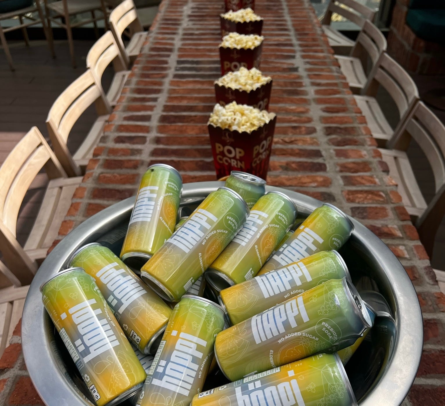 A bowl filled with cans of häppi prebiotic soda is placed on a long brick table, lined with popcorn buckets, creating a setup perfect for a movie night or casual gathering.