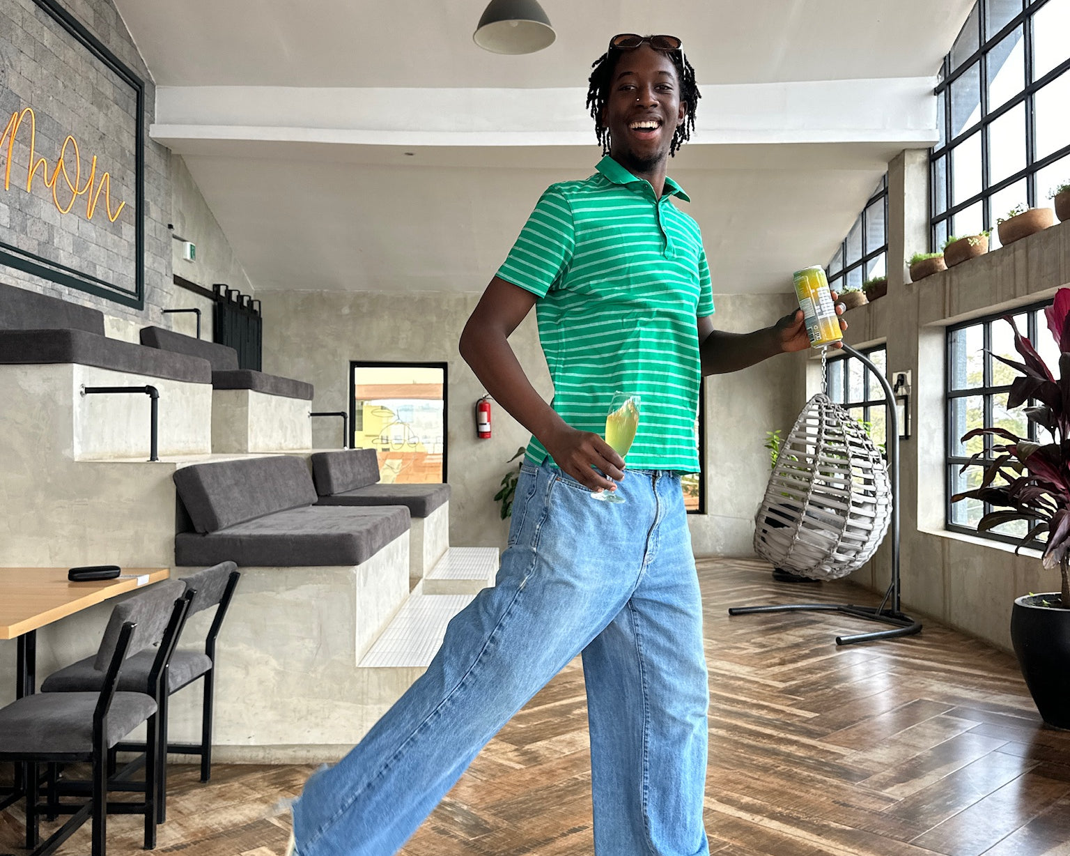 A cheerful person in a green-striped shirt and blue jeans holds a drink can, smiling mid-stride in a modern, well-lit café with large windows and cozy seating.