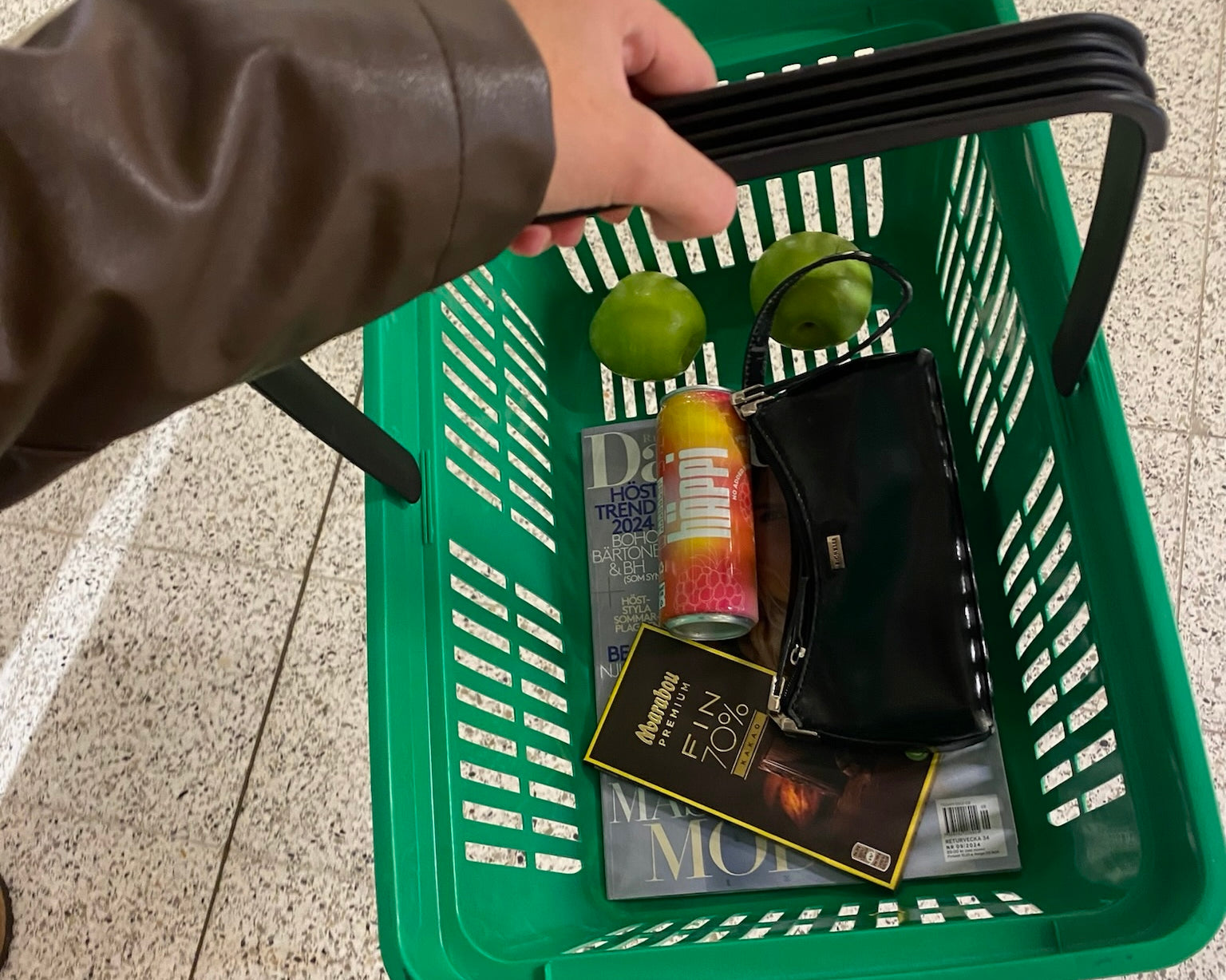 A green shopping basket featuring a hÄppi prebiotic soda, fresh green apples, a magazine, dark chocolate, and a black purse. 