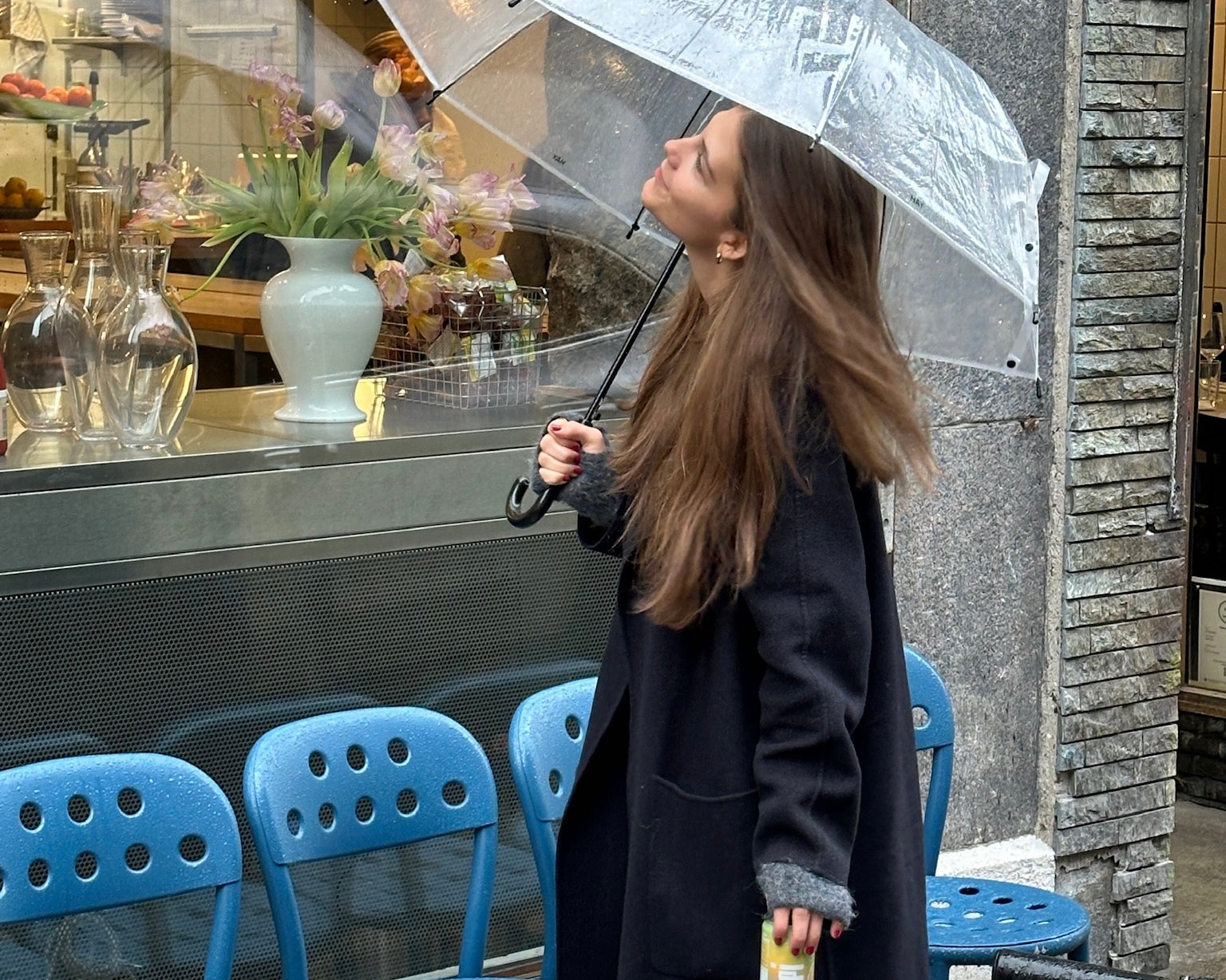 A woman enjoys a refreshing prebiotic soda on a rainy day, holding a clear umbrella and admiring her surroundings in a chic, urban setting