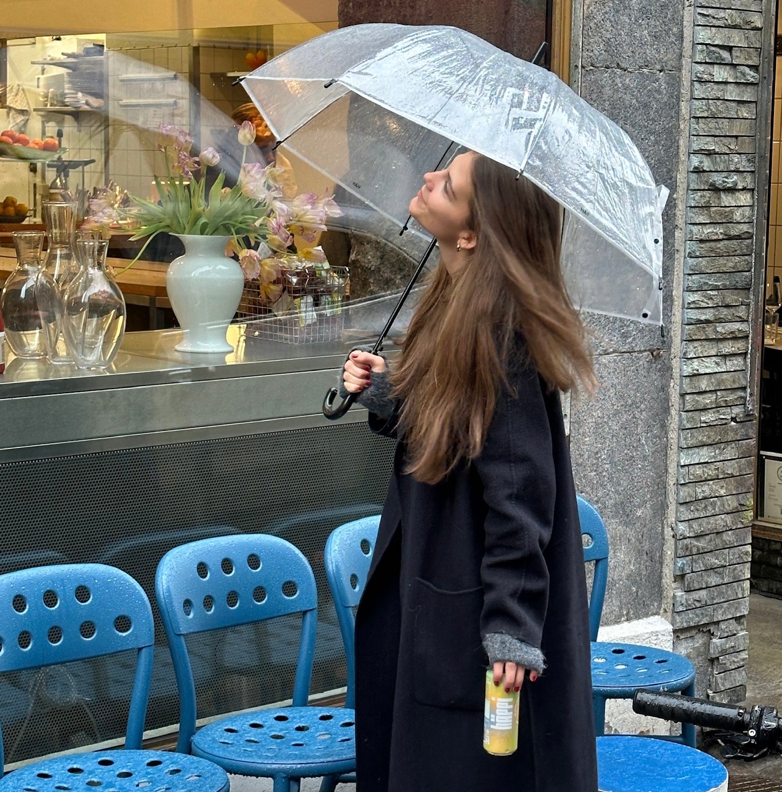 A woman enjoys a refreshing prebiotic soda on a rainy day, holding a clear umbrella and admiring her surroundings in a chic, urban setting