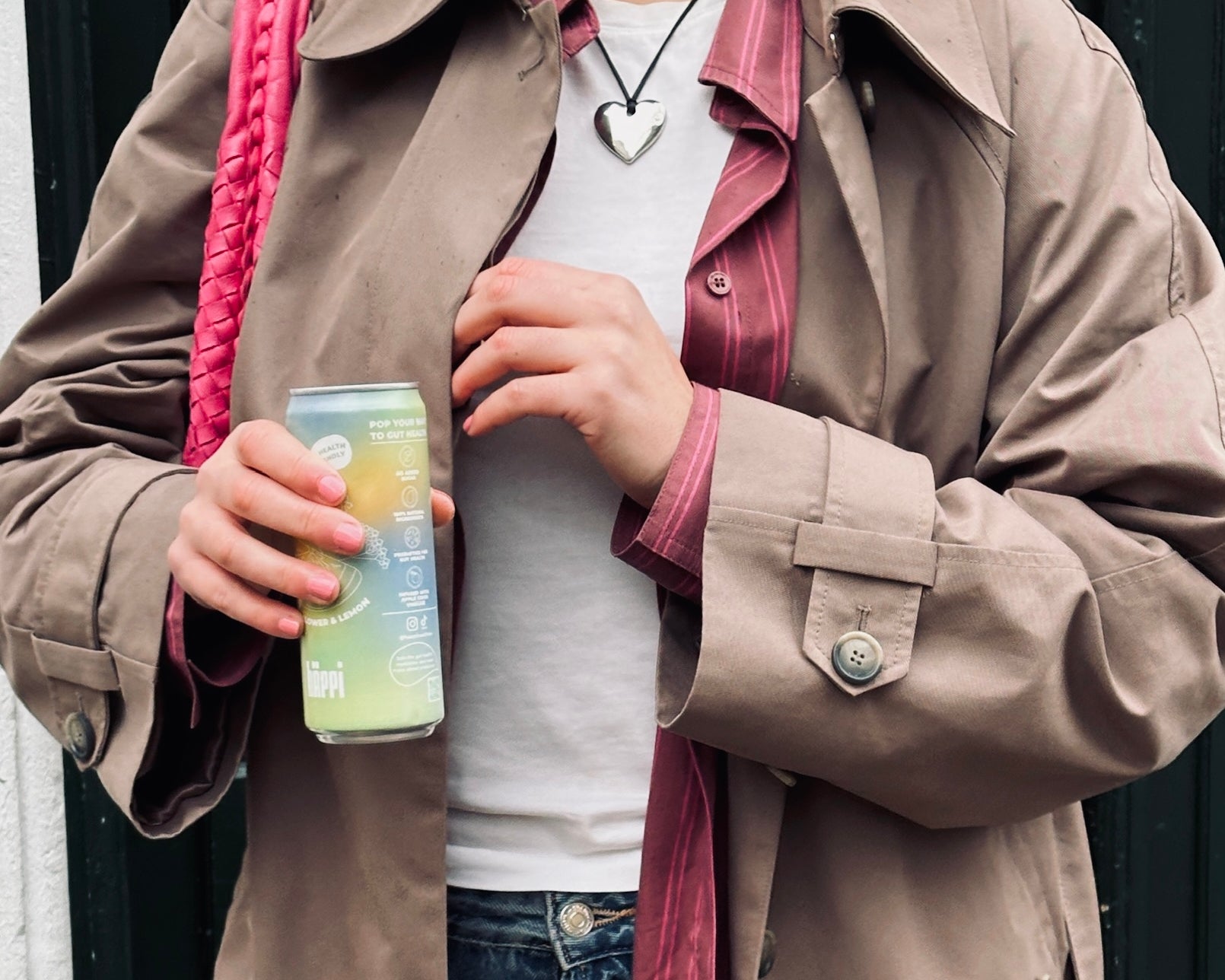 A person wearing a beige trench coat and a pink-striped shirt holds a can of hÄppi soda 