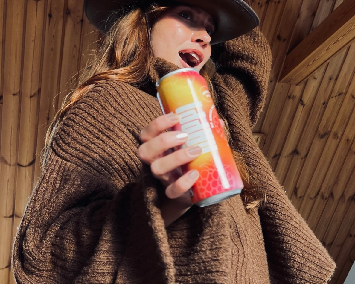 A woman in a cozy brown sweater and hat happily holds a vibrant hÄppi prebiotic soda can. The warm, rustic setting highlights the natural and wholesome vibe of the drink, perfect for health-conscious enjoyment.