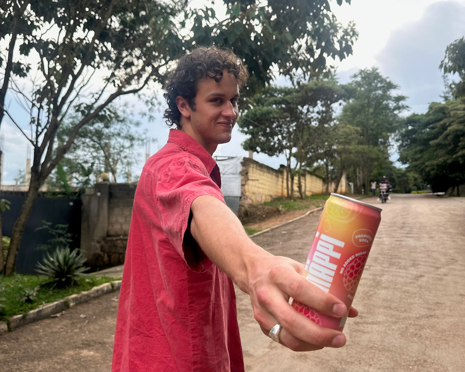 A young man in a red shirt offers a can of hÄppi soda while standing on a tree-lined street, smiling casually. The image conveys a relaxed, outdoor vibe.