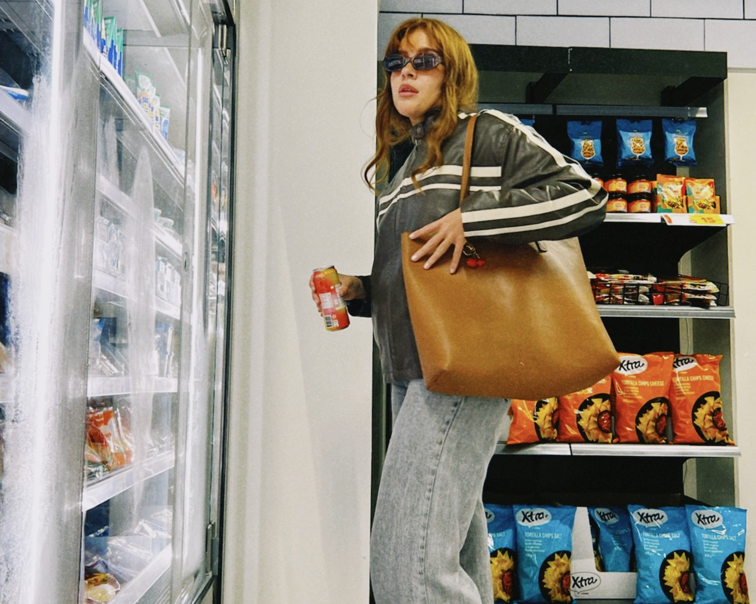 A stylish woman in sunglasses and a leather jacket holds a can of hÄppi soda while browsing a grocery store. She carries a large brown tote bag and stands in front of the freezer section, showcasing a trendy, casual look.